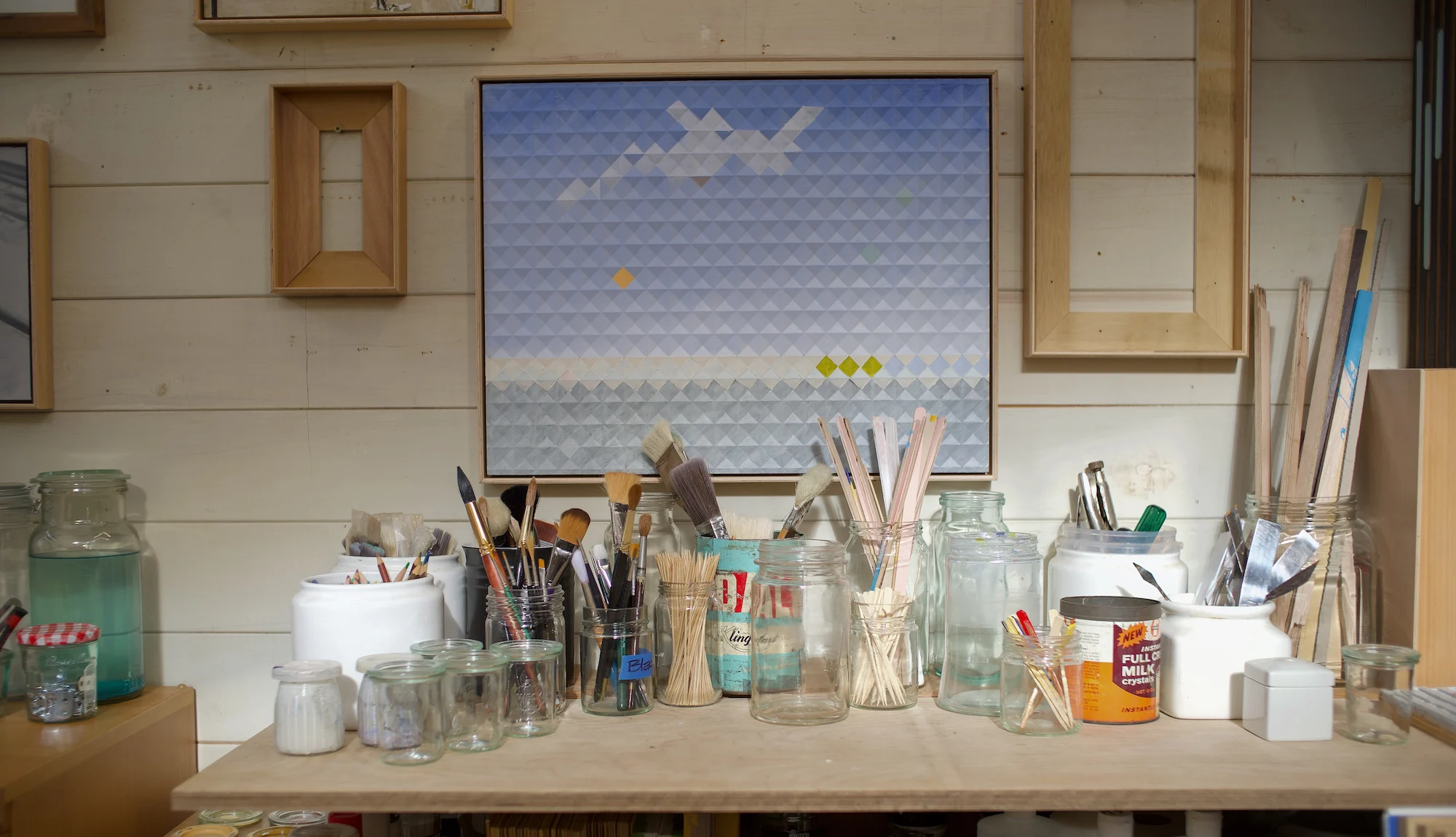 Shelf of artist's materials and containers with paintings and picture frames on wall behind.